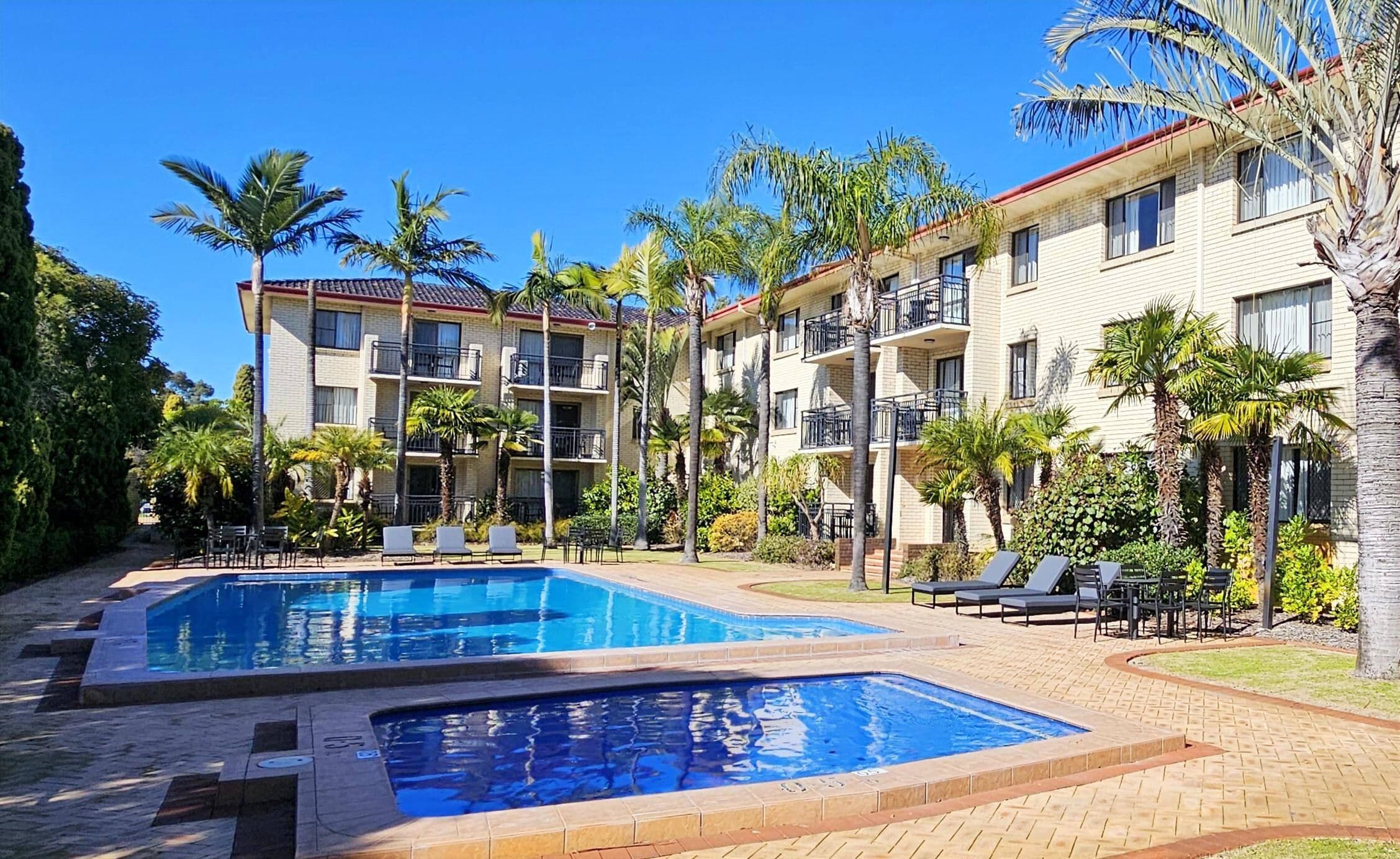A hotel with two swimming pools, surrounded by palm trees and a brick walkway.