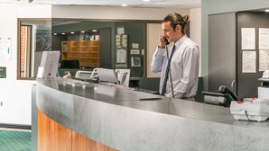 Ein Empfangsbereich mit einem an einem Telefon sprechenden Mitarbeiter, einer Überwachungskamera und einem Schalter mit Computer und Drucker.