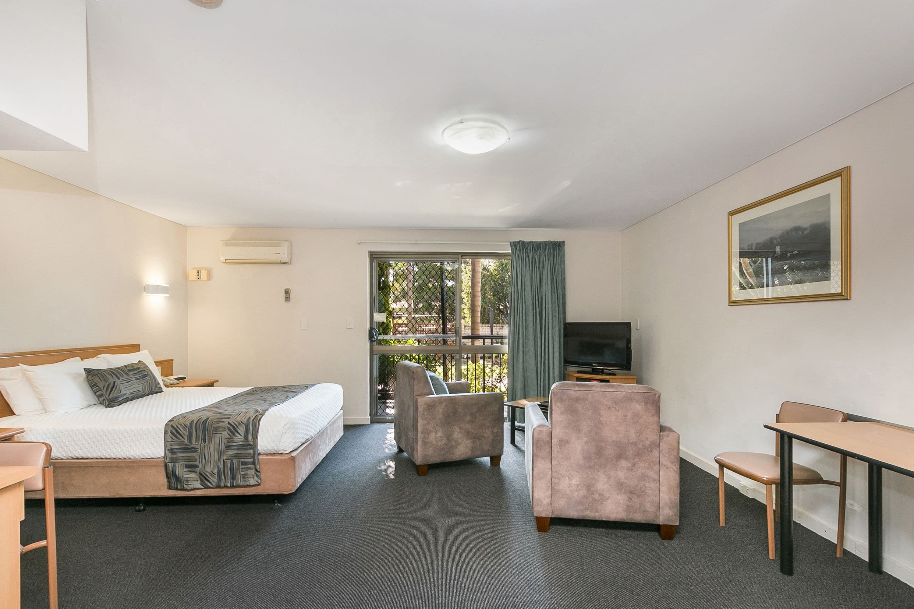 A hotel room with a bed, a desk, a chair, a TV, and a window with a view of trees.