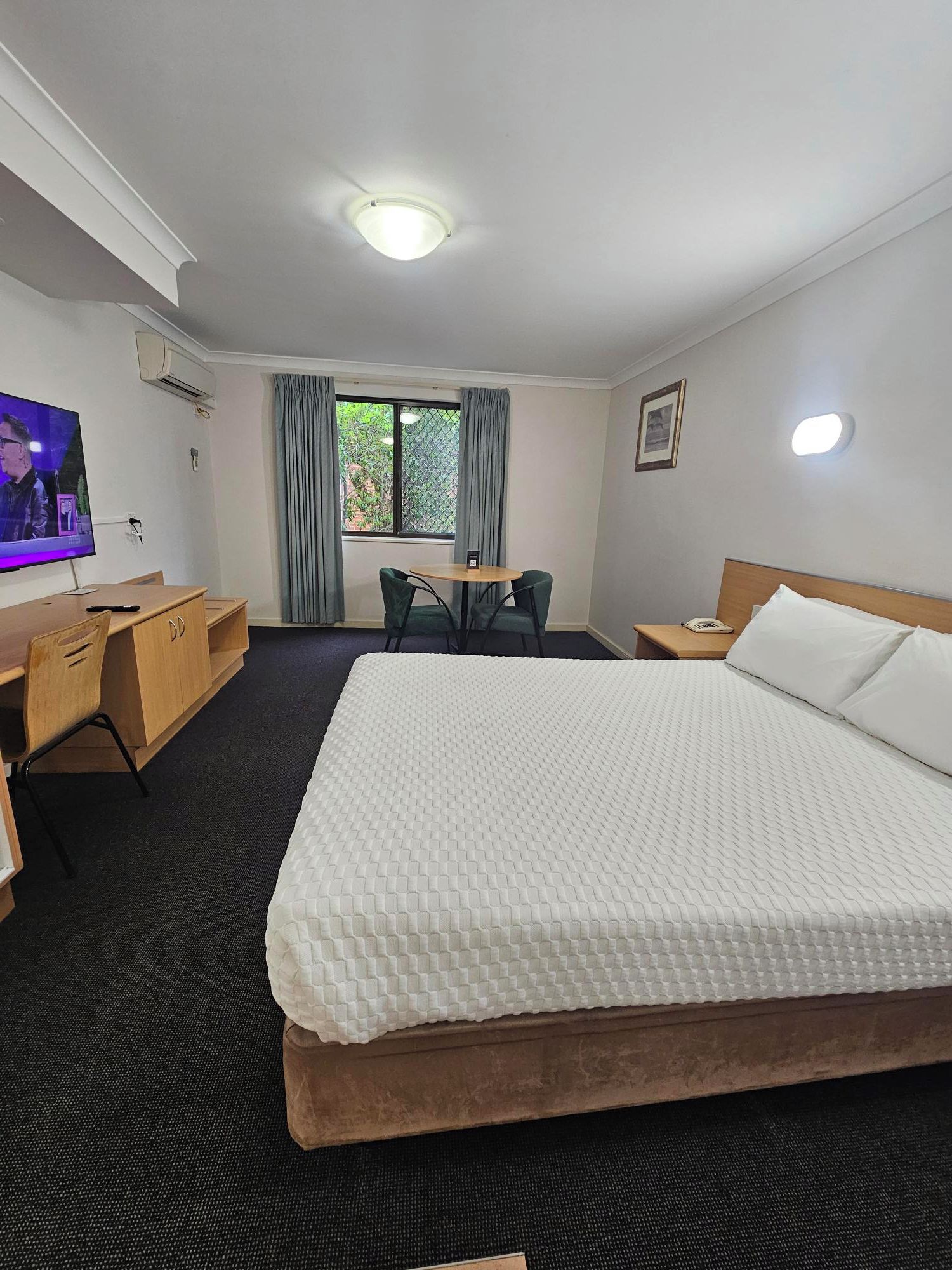 Ein Hotelzimmer mit einem großen Bett, einem Schreibtisch mit Stuhl, einem Fernseher und einem Fenster mit Vorhängen.