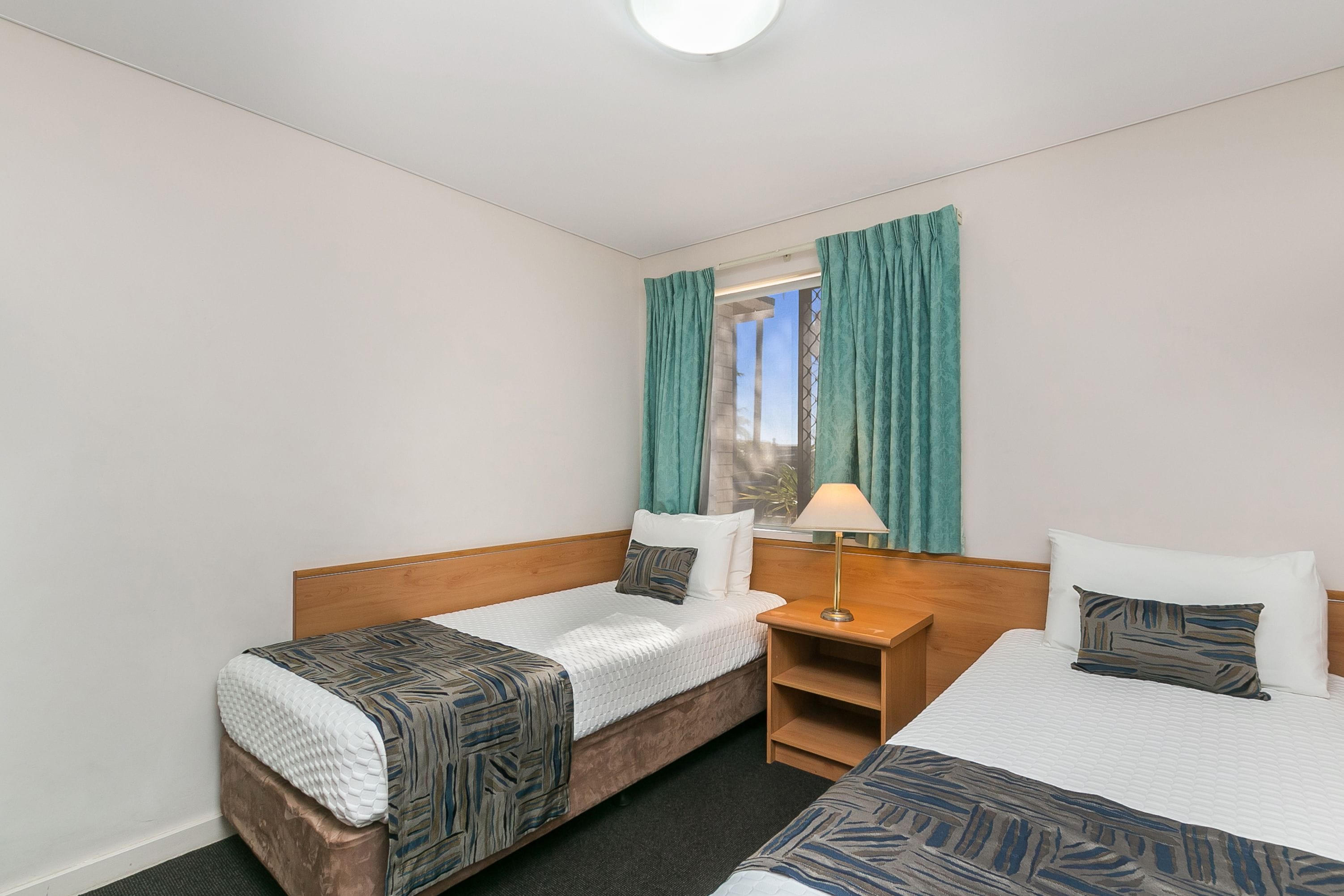Habitación de hotel con dos camas, cabecero de madera, mesita de noche y ventana con cortinas.