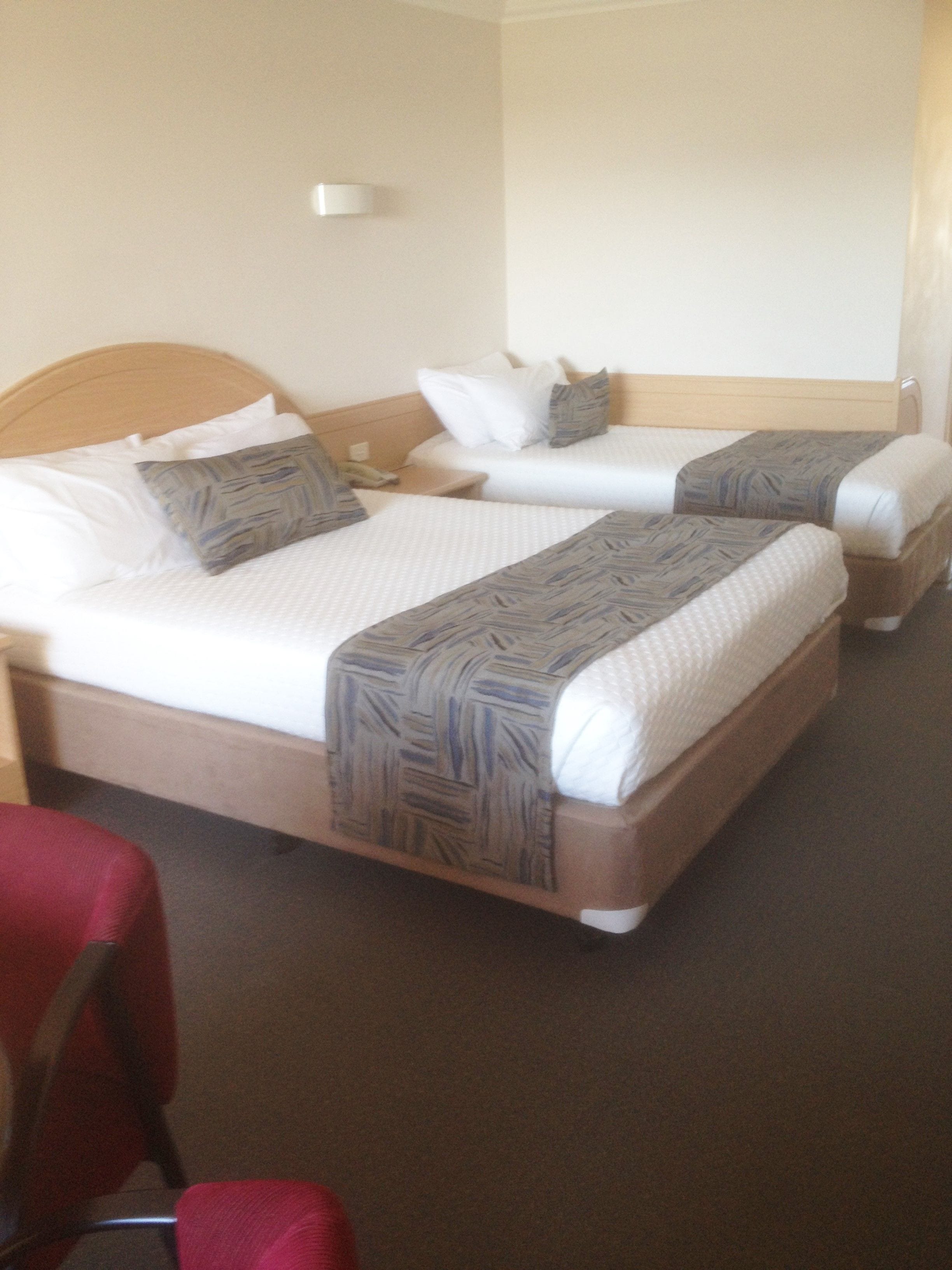 A hotel room with two beds, each with a patterned blanket and pillows, a wooden headboard, a bedside table with a lamp, and a red chair.