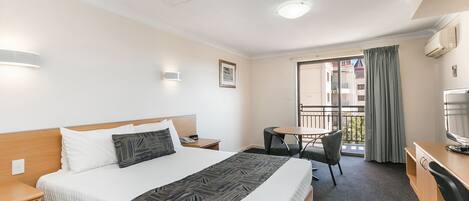 A hotel room with a large bed, a desk, a chair, a small table, and a balcony with a view of buildings.