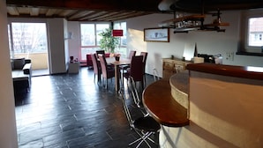 A spacious living area with a dining table, chairs, and a bar counter. There is a large window providing natural light and a view of the outside.