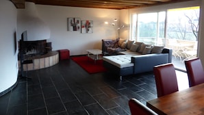 A modern living room with a fireplace, a sofa, a dining table, and a large window offering a view of the outdoors.