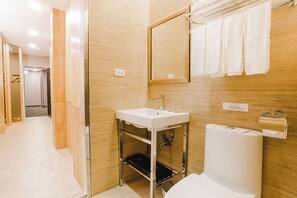 A modern bathroom with a white sink, mirror, and toilet. There is a towel rack above the toilet and a paper towel dispenser to the right.