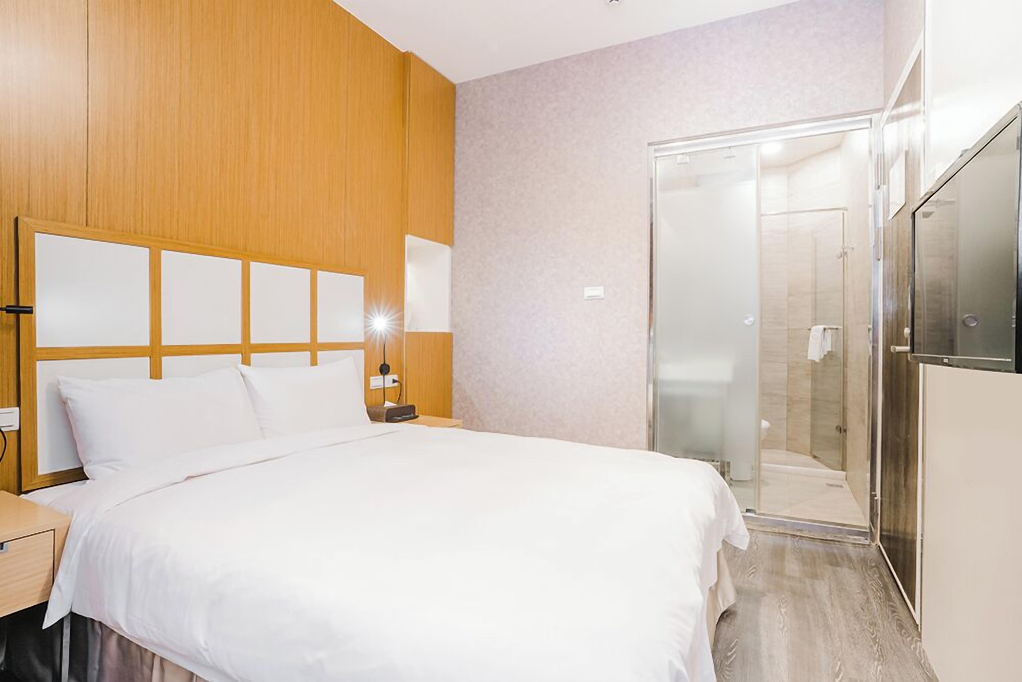 Una habitación de hotel con cama, mesita de noche, lámpara y zona de ducha con paredes de vidrio.