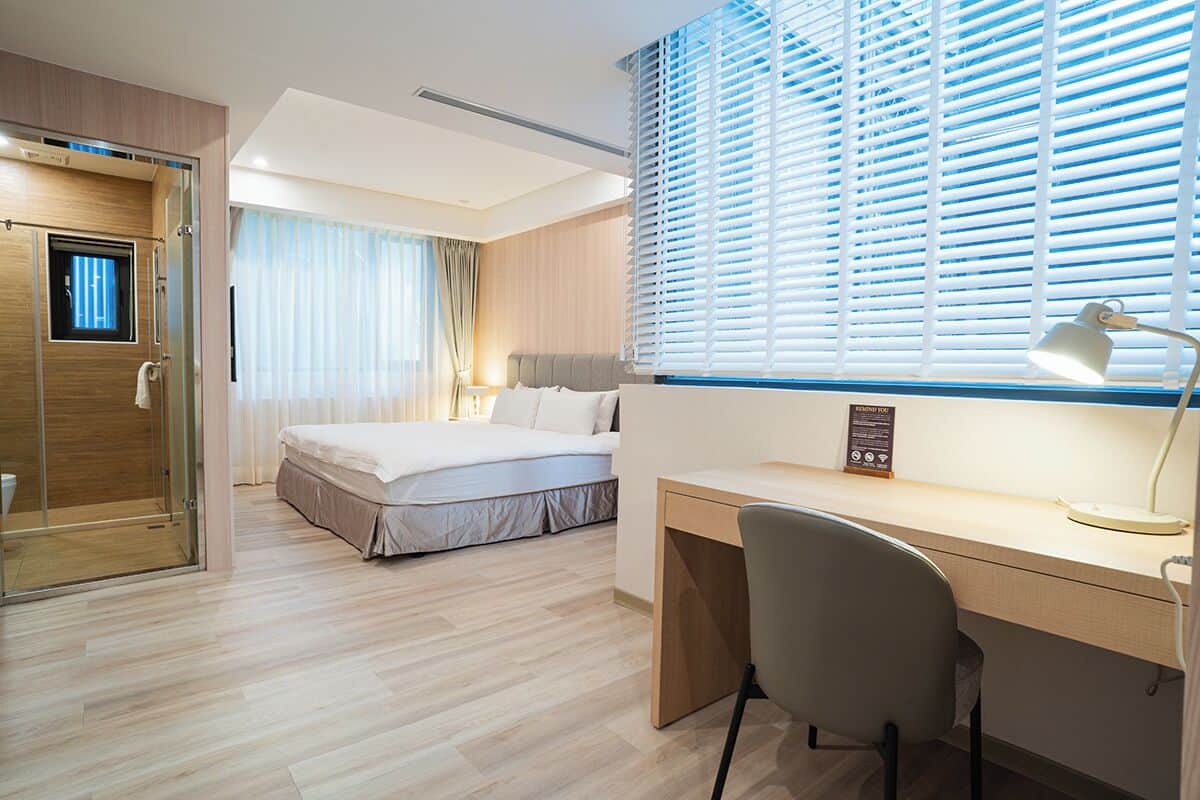 Ein modernes Hotelzimmer mit Bett, Schreibtisch, Stuhl und einem Fenster mit Jalousien.