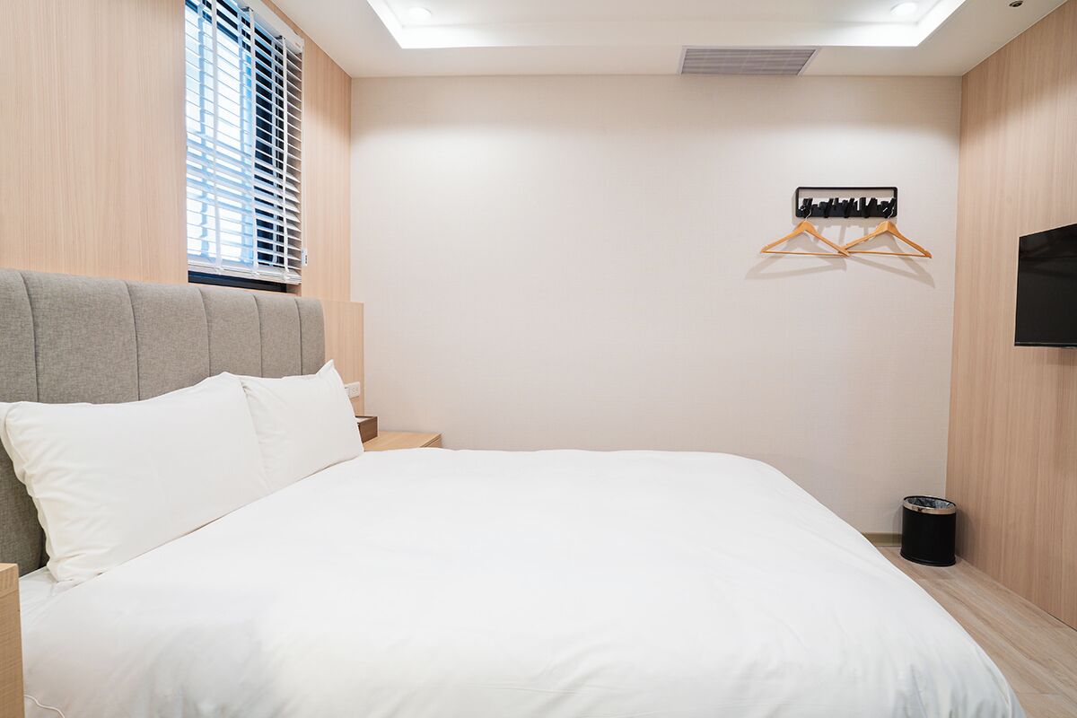 A bedroom with a bed, a wall-mounted TV, a window with blinds, and a coat rack.