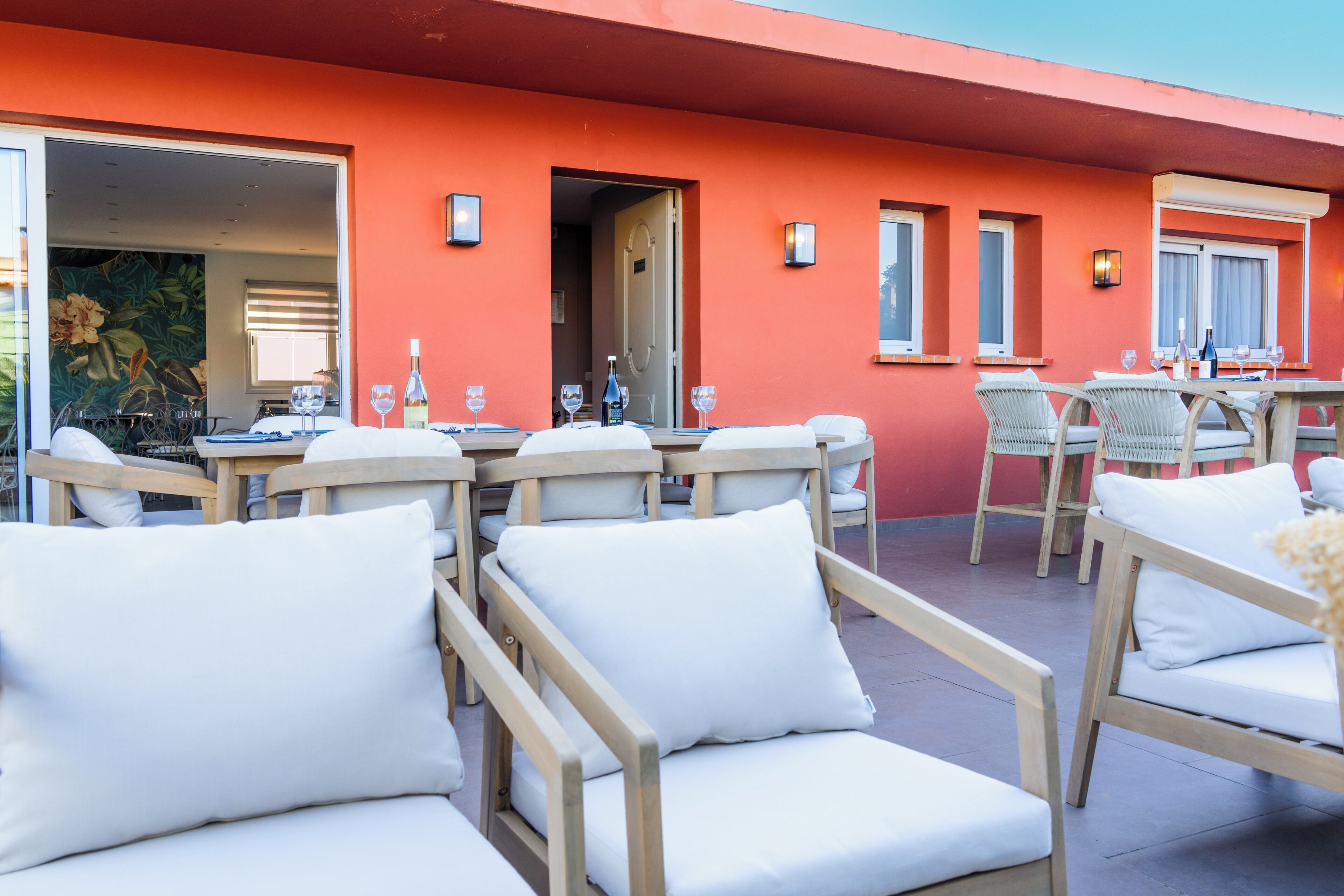 An outdoor seating area with white cushioned chairs and tables, a red wall, and a door leading inside.