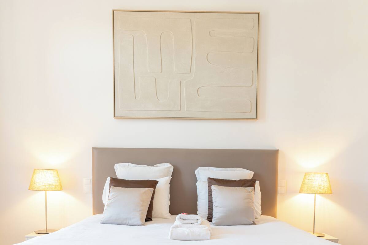 A neatly made bed with white bedding, two bedside lamps, and a large abstract artwork on the wall.