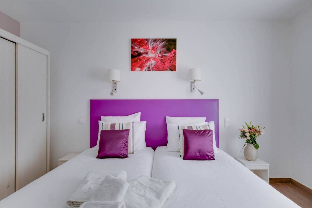 A double bed with white and purple bedding, a pink headboard, and a wall-mounted artwork above.