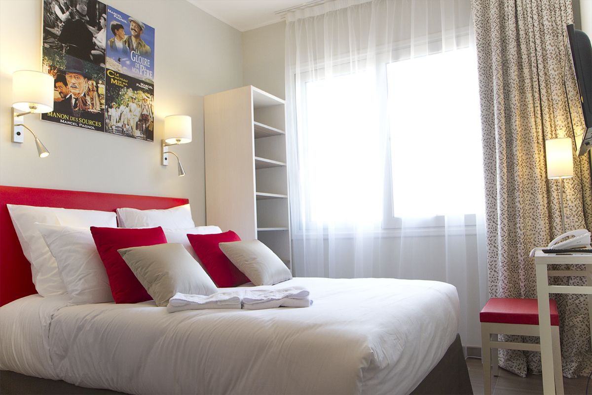 A hotel room with a bed, red and white pillows, a white headboard, a wall-mounted TV, a telephone, and a window with sheer curtains.