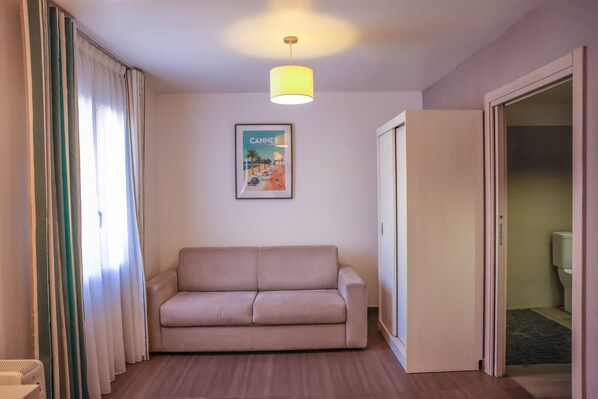 Una habitación con un sofá color beige, un cartel de Cannes, una puerta que da al baño y una lámpara en el techo.