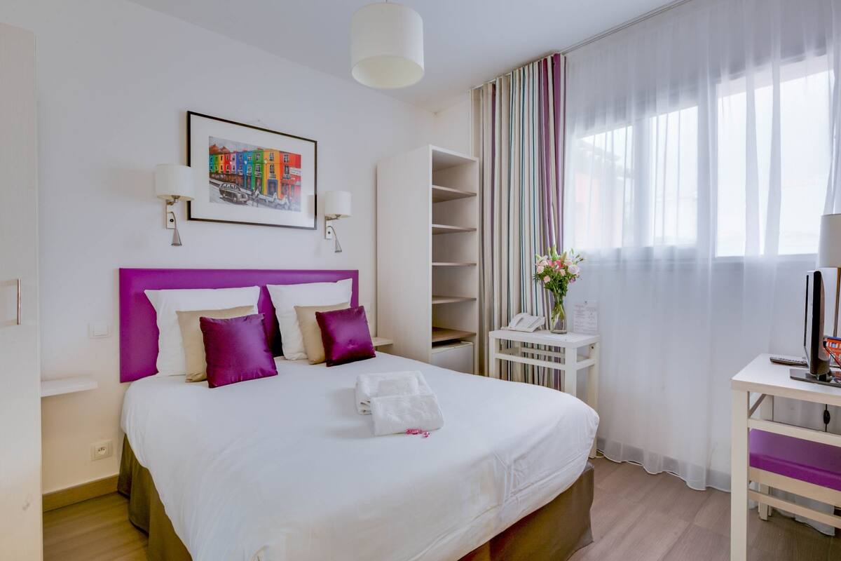 A hotel room with a large bed, a bedside table with a lamp, a small desk with a chair, and a window with sheer curtains.