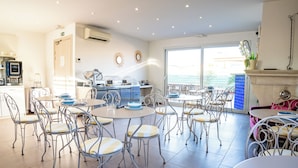 Uma área de refeições com mesas e cadeiras redondas, uma cozinha com eletrodomésticos e vista através de uma porta de vidro.