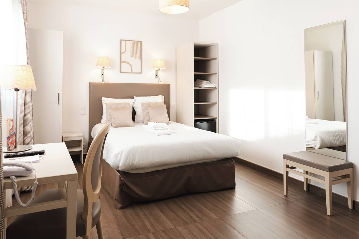 A hotel room with a bed, bedside tables, a desk, a chair, a wardrobe, and a mirror.