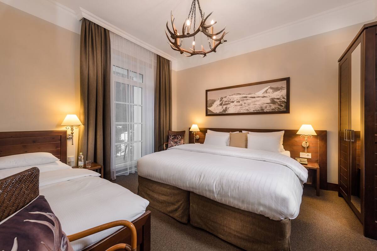 A hotel room with a large bed, two bedside tables with lamps, a chandelier, a painting on the wall, and a wardrobe.