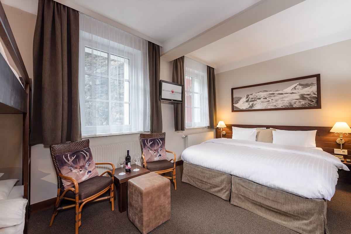 A hotel room with a bed, two chairs, a small table with a bottle and glasses, a TV, and a painting on the wall.