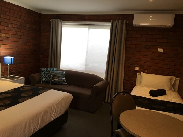 A hotel room with a brick wall, a large window with curtains, a sofa, a small table, and two beds.