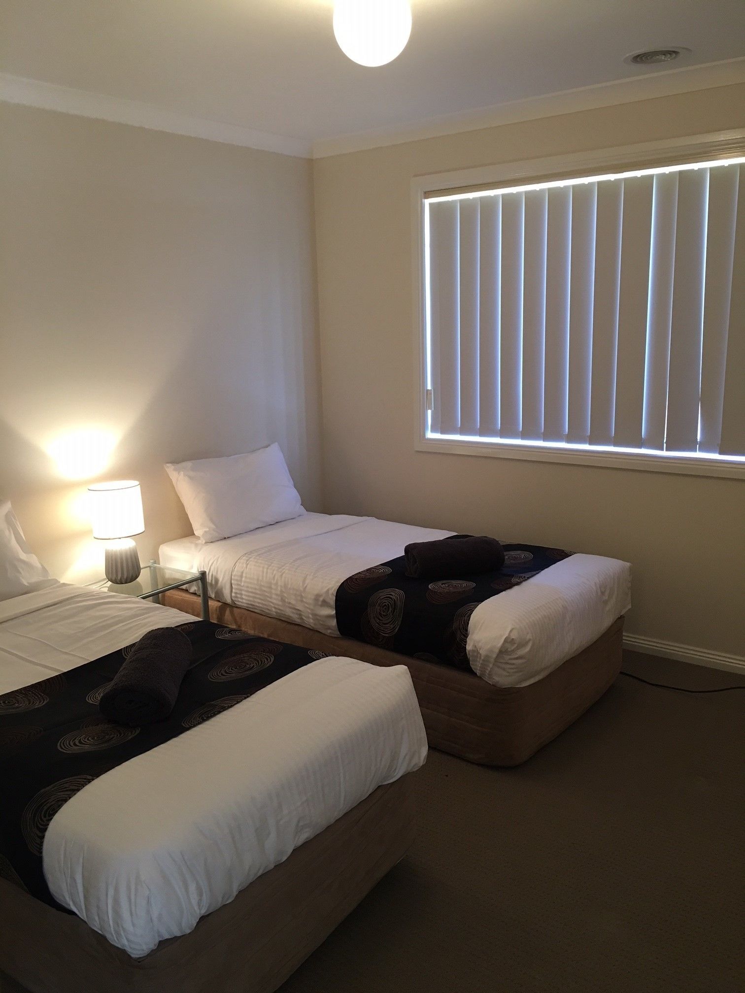 A bedroom with two beds, a nightstand, a lamp, and a window with blinds.