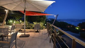 Une terrasse extérieure avec des tables et des chaises, un parasol blanc et une vue sur la mer et les collines.