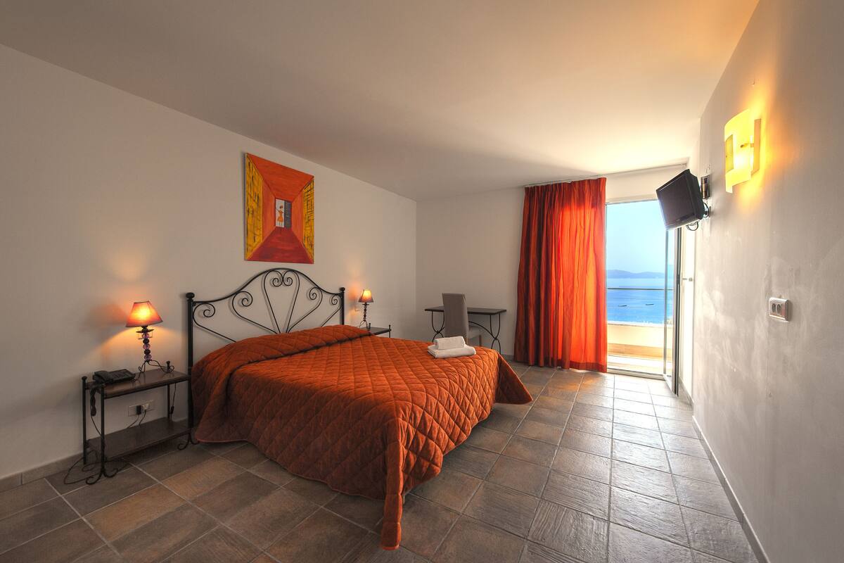Ein Schlafzimmer mit Bett, Nachttisch, Lampe, Gemälde, Sessel, Tisch und Meerblick durch ein Fenster.