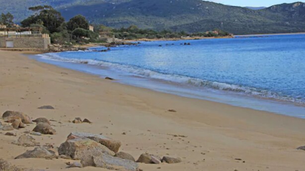 Une plage bordée d’un mur de pierres, de formations rocheuses et dominée par des montagnes en arrière-plan.