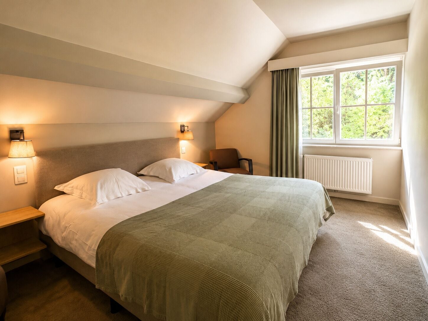 A hotel room with a bed, two bedside lamps, a chair, a window with curtains, and a radiator.