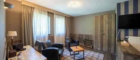 A hotel room with a wooden desk, two black chairs, a plaid rug, a television, and a fireplace.