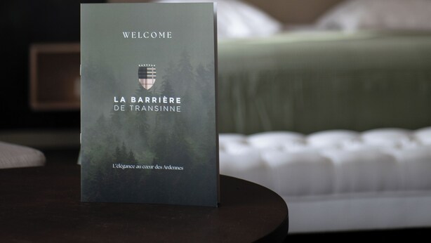 A welcome sign with text in a foreign language, a bed with white bedding, and a dark-toned headboard.