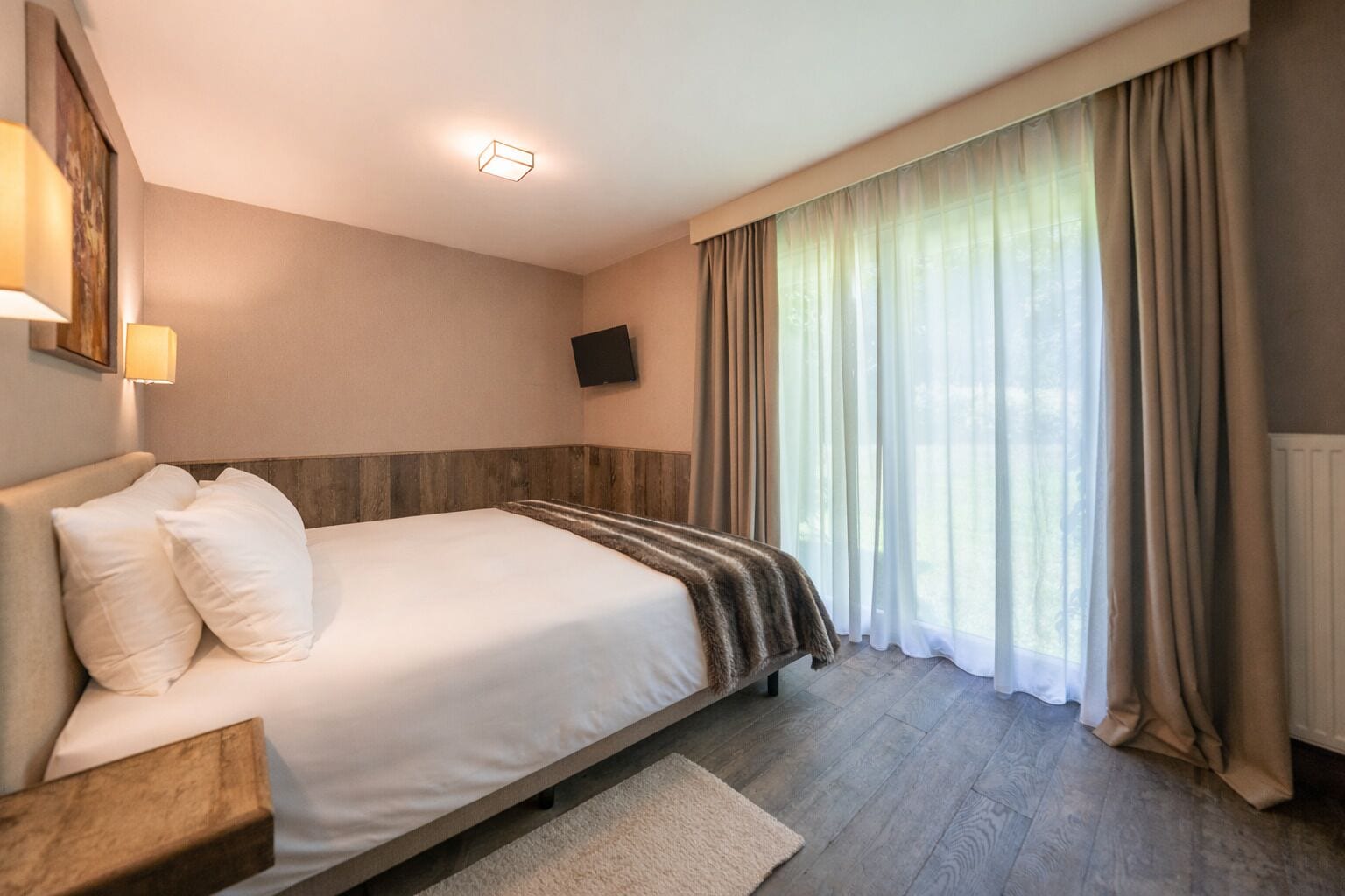 A hotel room with a bed, a TV mounted on the wall, and a window with curtains.