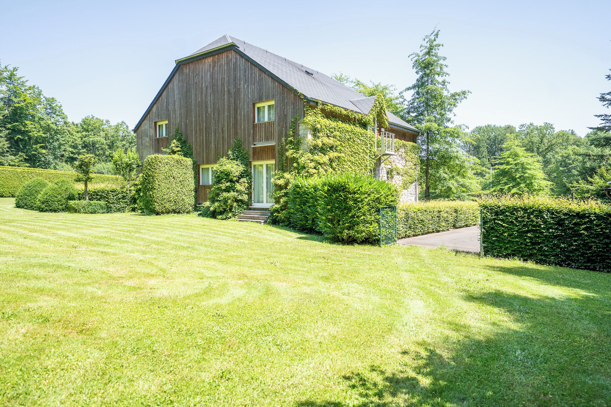 Een groot houten huis met schuin dak, omgeven door verzorgde gazons en hagen.