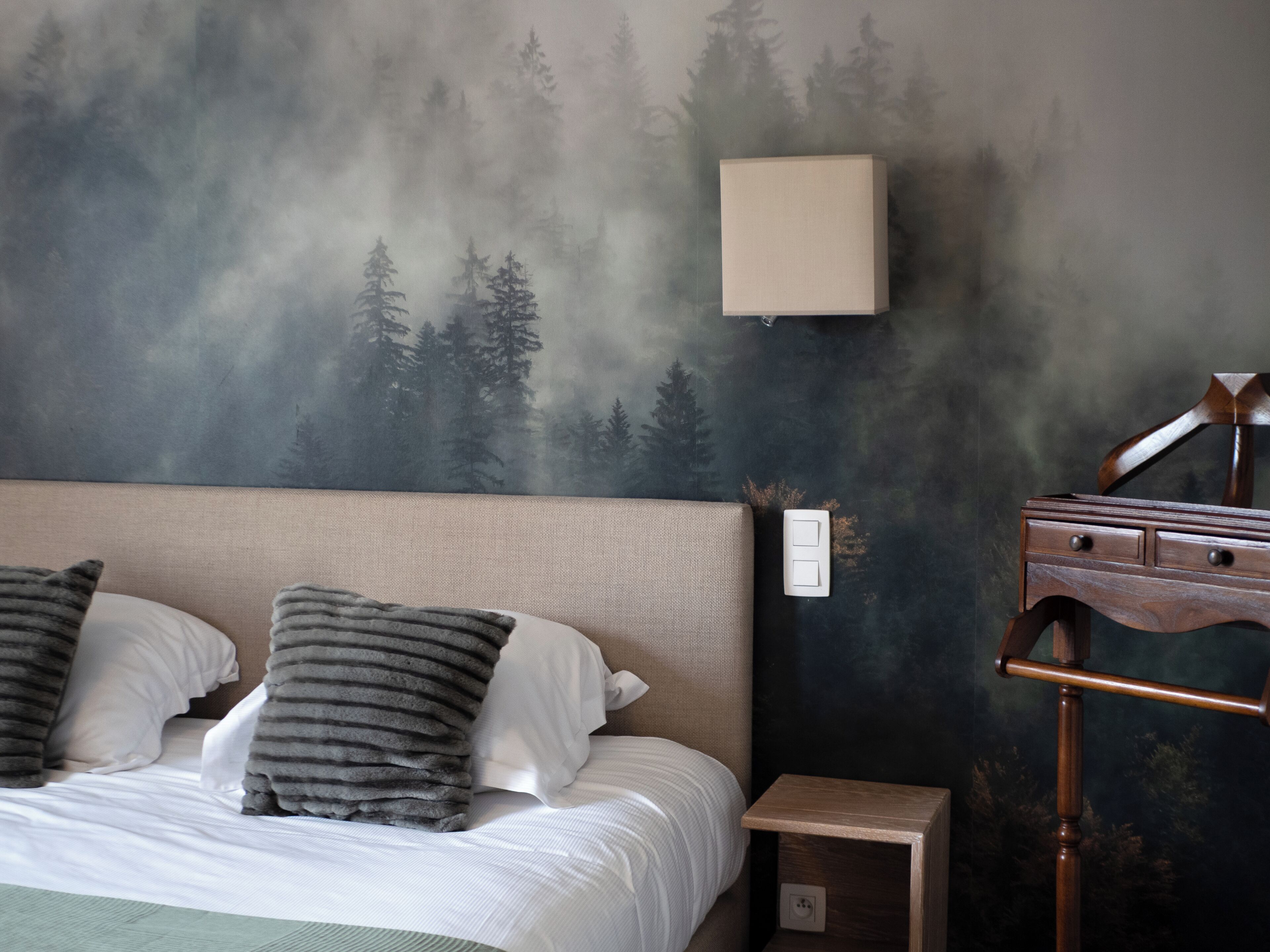 A bedroom with a bed, pillows, a nightstand, a lamp, and a wall with a forest mural.