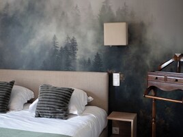 Un dormitorio con cama, almohadas, una mesita de noche, una lámpara y un mural en la pared con un bosque.