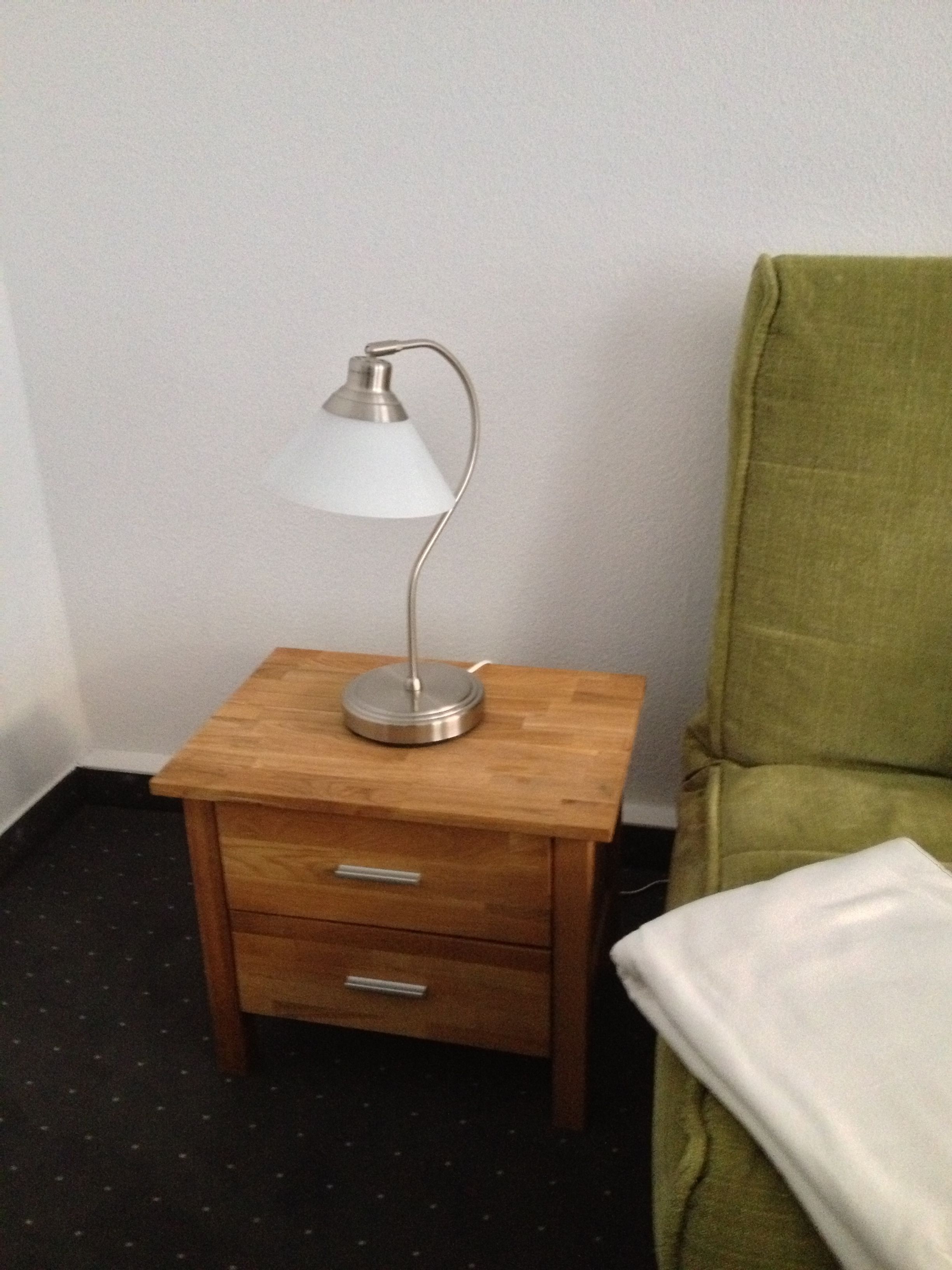 Una mesita de noche de madera pequeña con una lámpara y dos cajones, ubicada al lado de una silla verde con un cojín blanco.