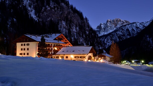 Un albergue de montaña cubierto de nieve con varias edificaciones, incluyendo un edificio principal y varias cabañas más pequeñas, rodeado de árboles y montañas nevadas.