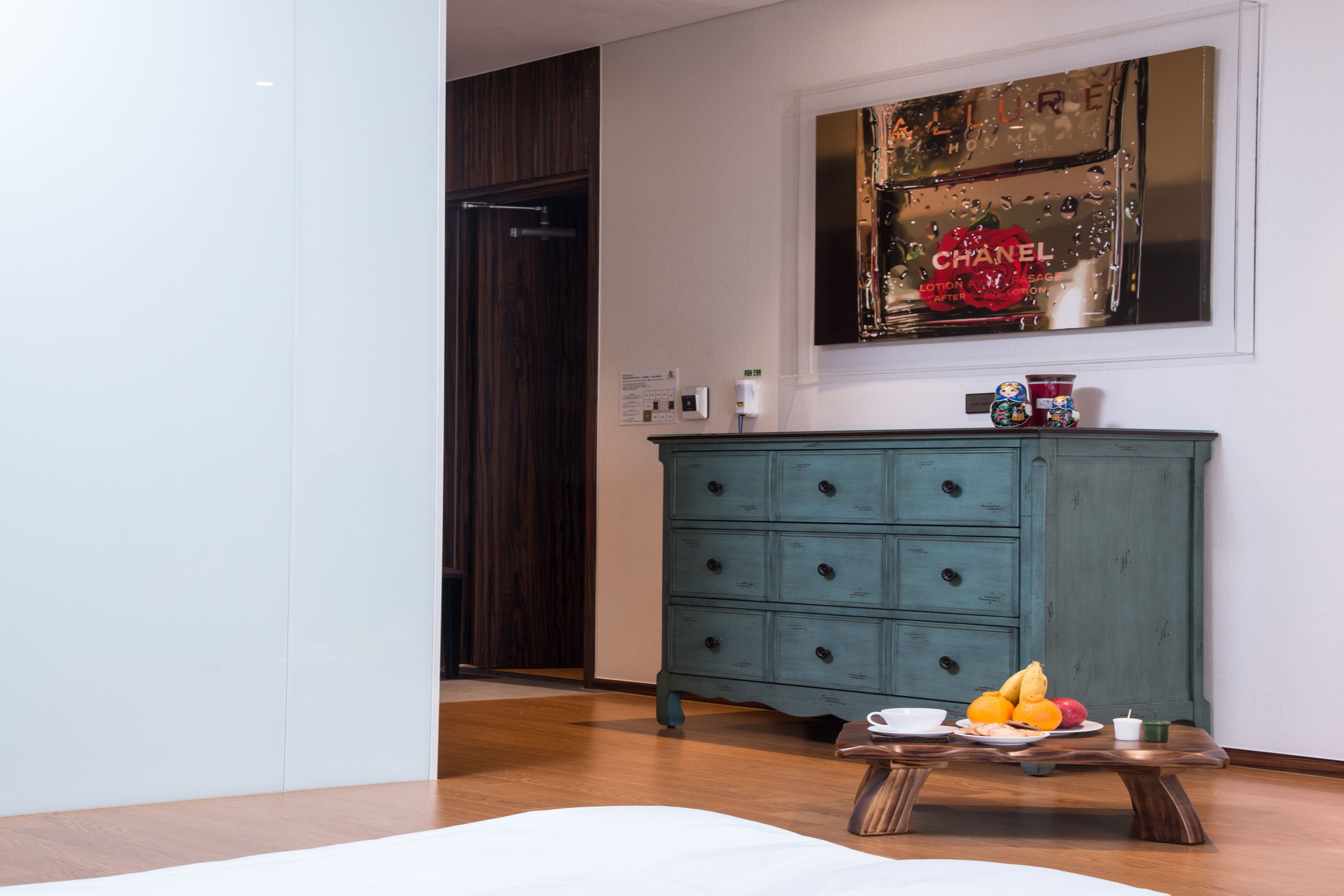 A room with a wooden floor, a teal dresser, a table with fruits and a cup, and a wall with a framed picture.