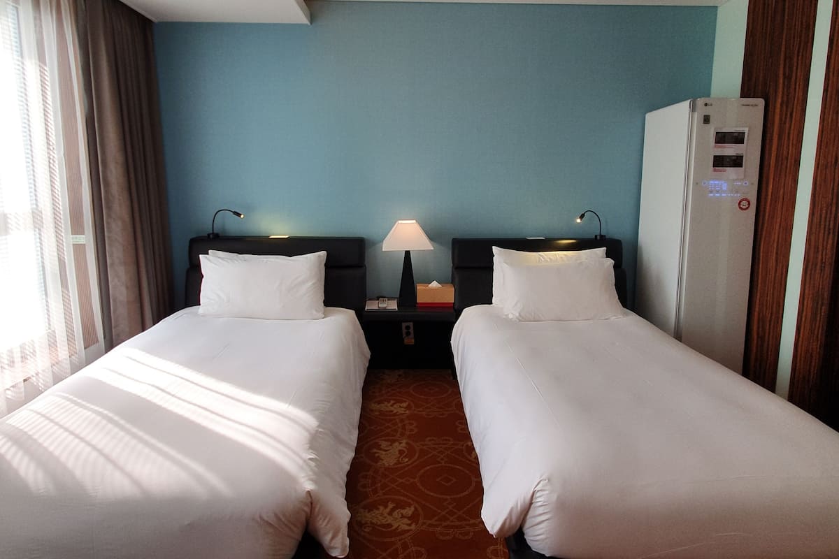 Ein Hotelzimmer mit zwei Einzelbetten, einem Nachttisch mit Lampe, einer Minibar und einem Kühlschrank.