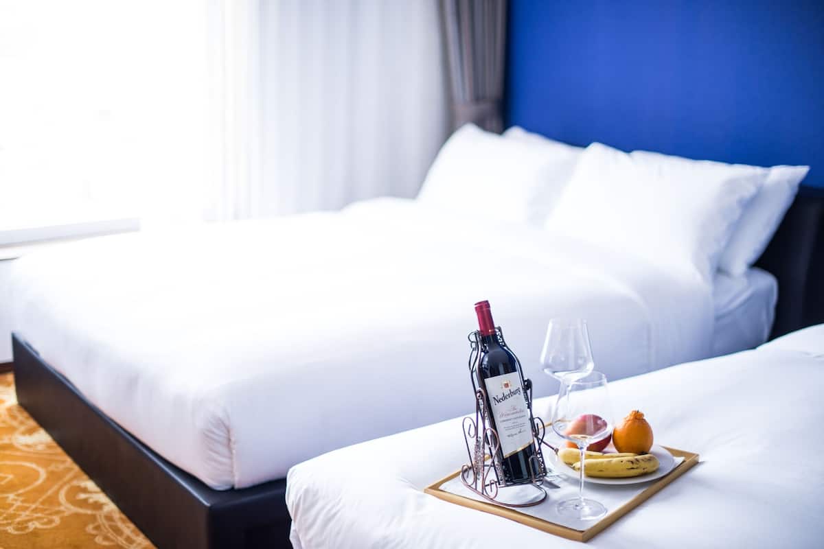 Ein Hotelzimmer mit einem ordentlich bezogenen Bett, einem Tablett mit einer Weinflasche, einem Glas und Obst.