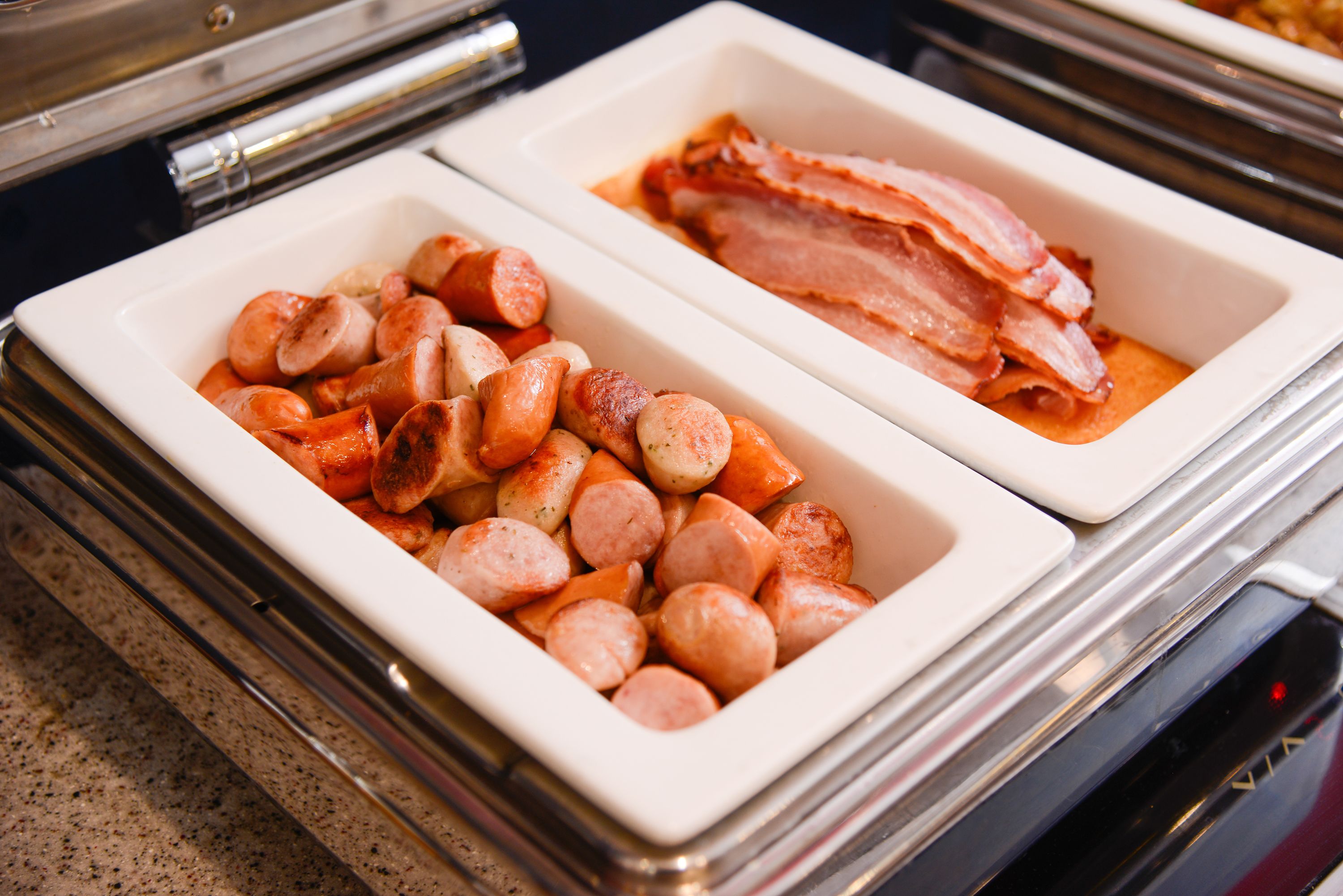 Two white rectangular containers on a countertop, one filled with small sausages and the other with bacon strips.