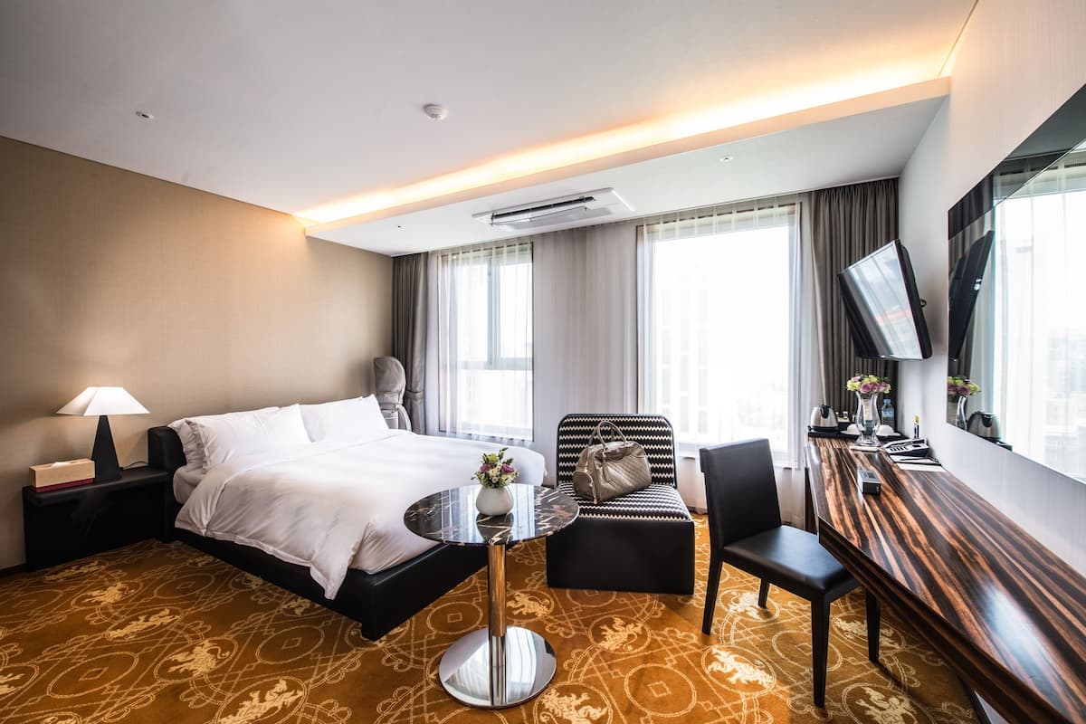 Ein Hotelzimmer mit Bett, Schreibtisch, Stuhl, einem kleinen Tisch, einer Lampe, einem Fernseher und einem Fenster mit Vorhängen.