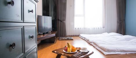 Habitación de hotel con piso de madera, cama, televisor sobre un pedestal, banca de madera con fruta y una taza, y un ventanal con cortinas.
