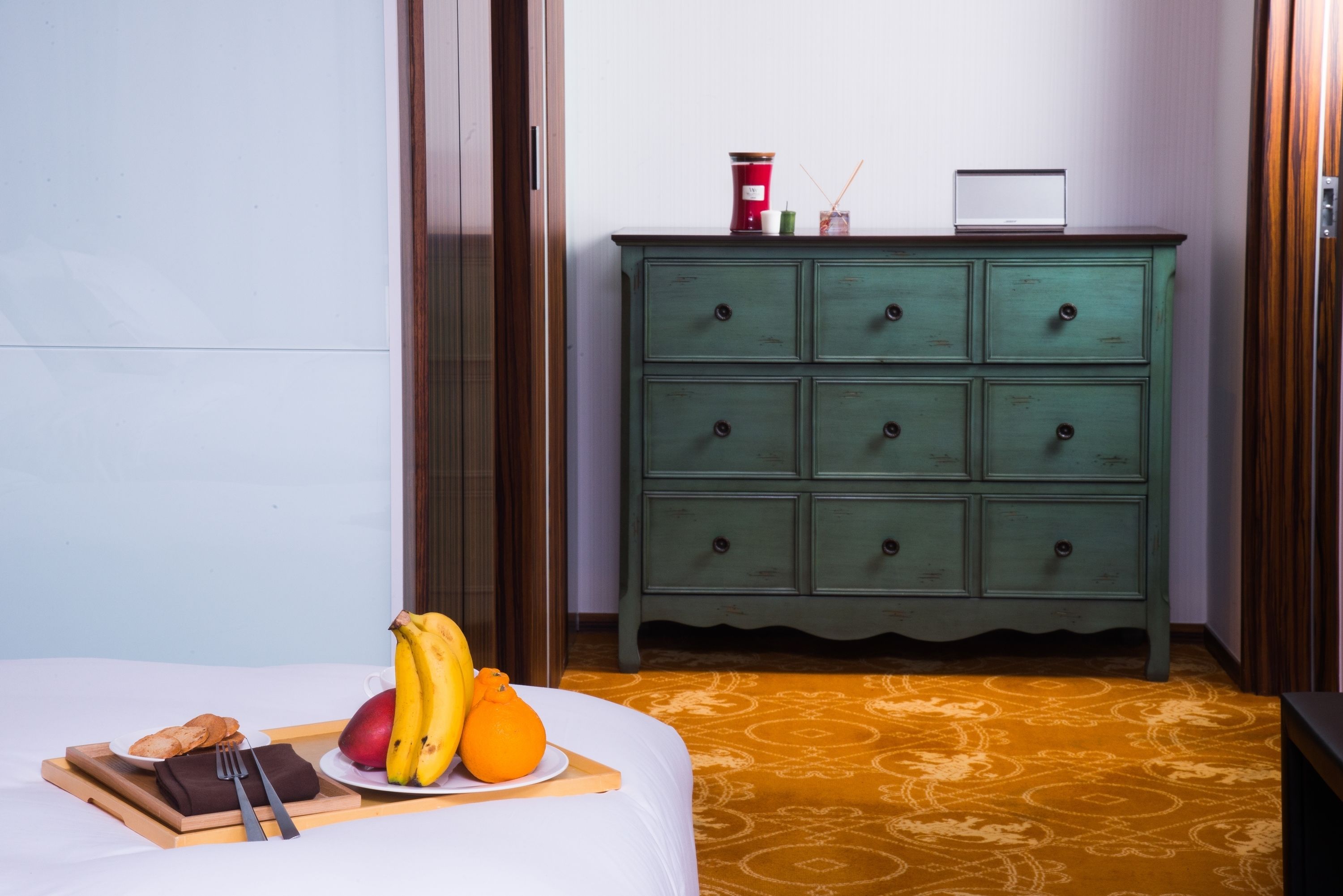 A hotel room with a bed, a green dresser, a tray with fruits and a cup, and a small table with a laptop.