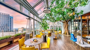 A modern indoor dining area with yellow chairs, a large tree, and a view of the cityscape through large windows. - Hotel Leo (Jeju City)