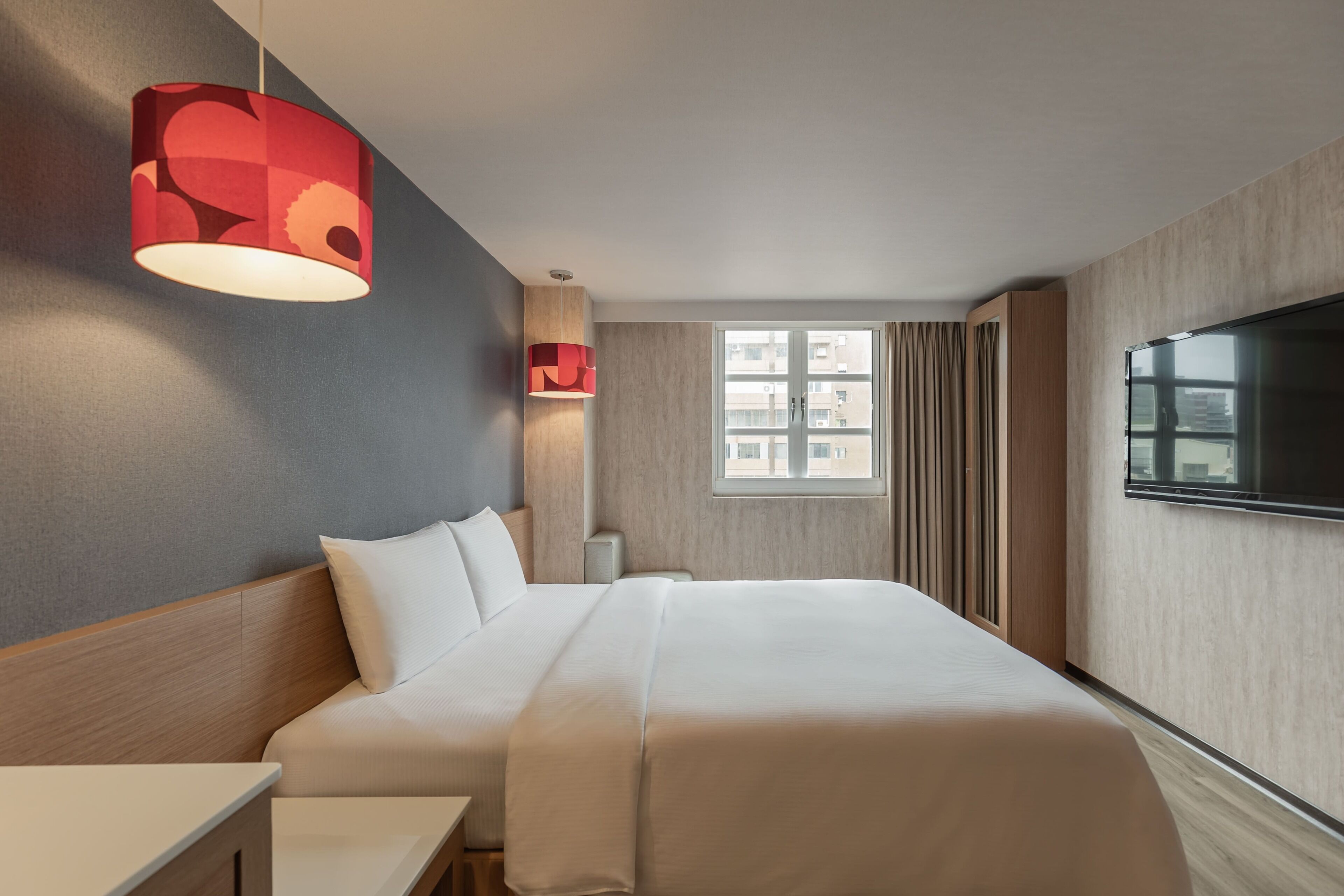 Una moderna camera d'albergo con un grande letto, una TV a parete, una finestra con tende e una particolare lampada a sospensione rossa.