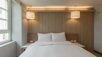 A modern hotel room with a wooden headboard, two bedside lamps, a large bed, and a window providing a view of the city.