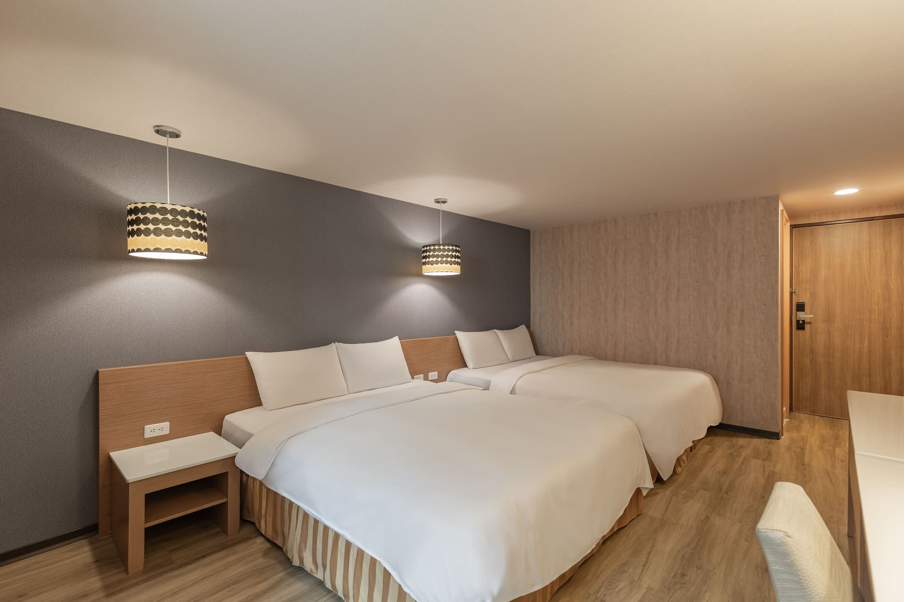 Habitación de hotel con dos camas, cabecero de madera, mesita de noche pequeña y dos lámparas colgantes.