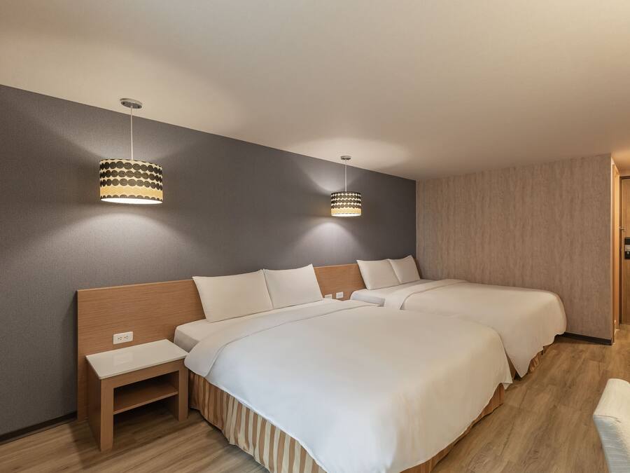 Habitación de hotel con dos camas, cabecero de madera, mesita de noche pequeña y dos lámparas colgantes.