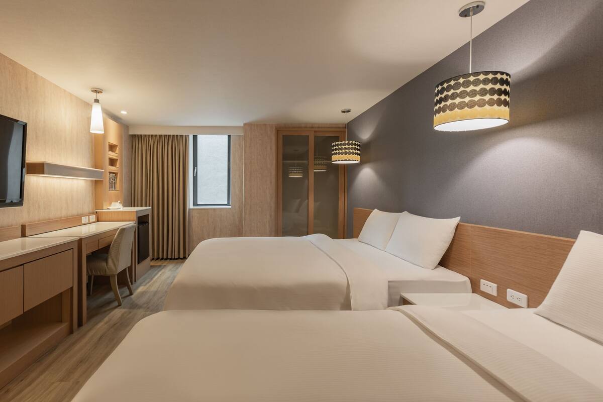 Ein Hotelzimmer mit zwei Betten, einem Schreibtisch, einem Sessel, einem Fernseher und einem großen Fenster mit Vorhängen.
